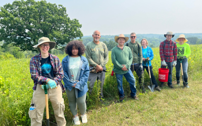 Smiling group of 8 volunteers standing in a prairie with parsnip predator (shovel looking tool) after a volunteer workday