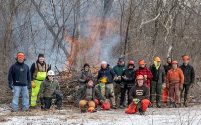 Crew of 15 volunteers and staff saying "cheese" while standing in front of a burn pile