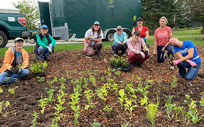 Volunteers kneeling in a garden, planting young plants with a green trailer and truck in the background.