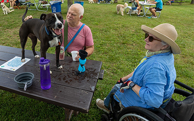 Two people at a picnic table with a dog on the table looking very happy.