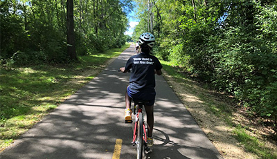 Teenager biking along a shady portion of the Lower Yahara River Trails. Trees on both side of the paved path.
