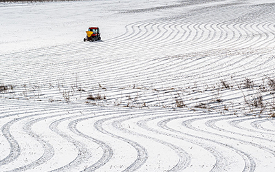 A small vehicle with a seed spreader attached driving on a snowy fields. Tire tracks show the path it has taken back and fourth. 