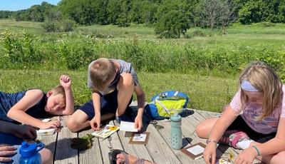 Kids sitting on a wooden platform pressing flowers. Prairie in the background.