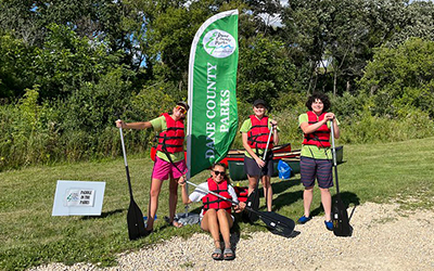 A group of people with canoe paddles wearing red personal flotation devices standing next to a green "Dane County Parks" flag