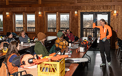 Person in orange jacket giving a chainsaw safety demonstration to a group seated at tables inside a wooden building.