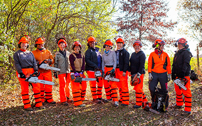 A group of people wearing orange safety gear and helmets outdoors holding chainsaws on a sunny day with trees in the background.
