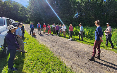 A group of people standing in a semi-circle outdoors on a sunny day near a grassy area and trees.