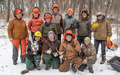 Group of people outdoors wearing safety gear and holding tools in a snowy forest setting.